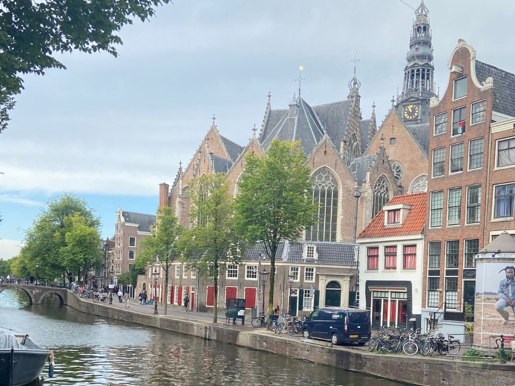 Ein Amsterdamer Grachtenkanal mit historischen Giebelhäusern und der Oude Kerk im Hintergrund; am Ufer parken zahlreiche Fahrräder.