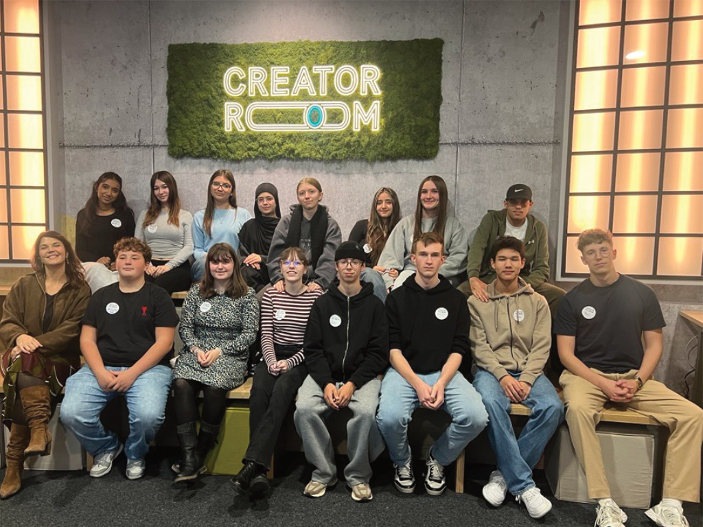 Eine Gruppe Jugendlicher sitzt mit einer erwachsenen Begleitung in zwei Reihen vor einer Moswand mit dem leuchtenden Schriftzug „CREATOR ROOM“.
