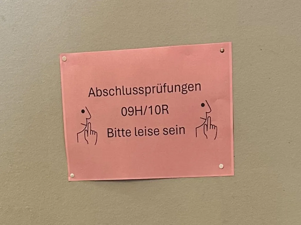 Das Foto zeigt einen ruhigen Schulflur einer Realschule. Im Mittelpunkt steht eine große, freistehende graue Stellwand, die fast den gesamten Blick versperrt. An der Stellwand hängt ein rosa Blatt Papier mit der Aufschrift:    „Abschlussprüfungen 09H/10R – Bitte leise sein“    Neben dem Text sind auf beiden Seiten Symbole von Fingern zu sehen, die zum Stillsein auffordern.    Der Flur ist modern gestaltet. Rechts sind Wände und Stützen aus hellem Holz zu sehen, darüber verläuft ein silbernes Lüftungsrohr unter der Decke. Im Hintergrund führt ein Gang zu mehreren Räumen mit geschlossenen Türen.