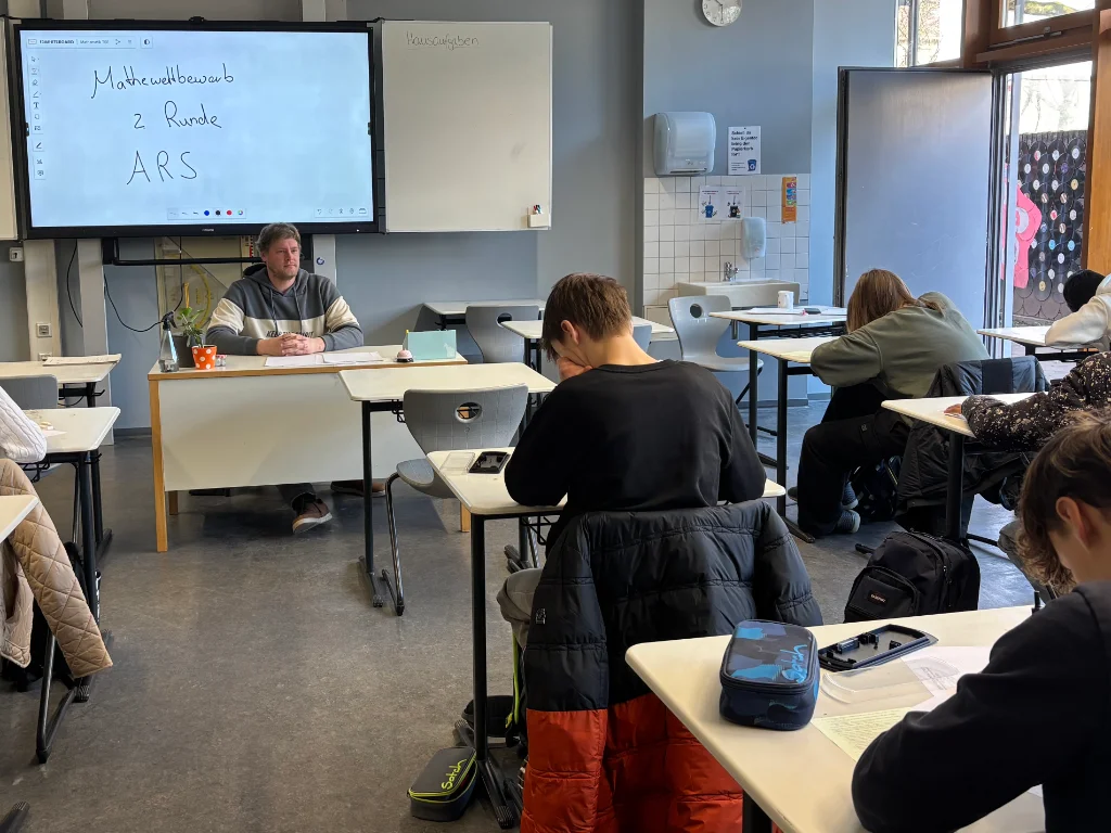 Jugendliche schreiben an Einzeltischen den Mathematikwettbewerb; vorne beaufsichtigt eine Lehrkraft, auf dem Smartboard steht „Mathewettbewerb 2. Runde ARS“.