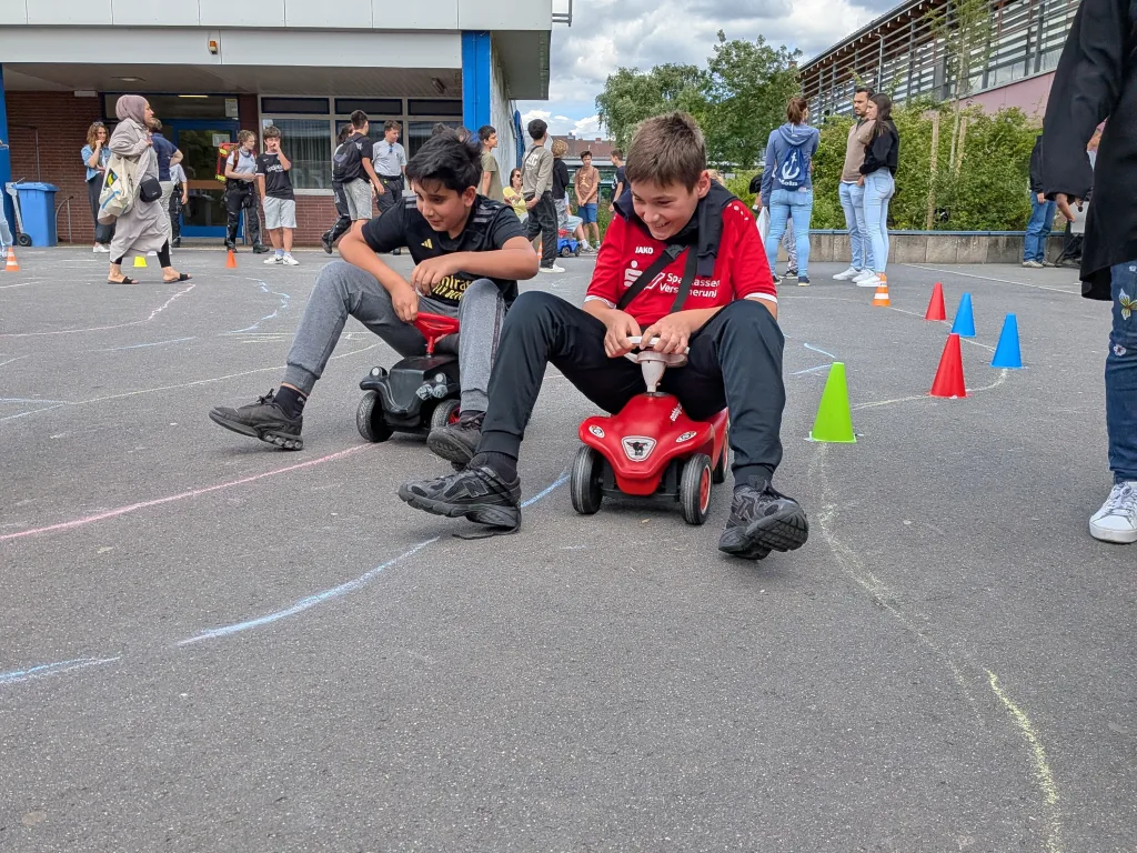Zwei Jugendliche fahren auf Bobbycars einen Parcours mit Hütchen.