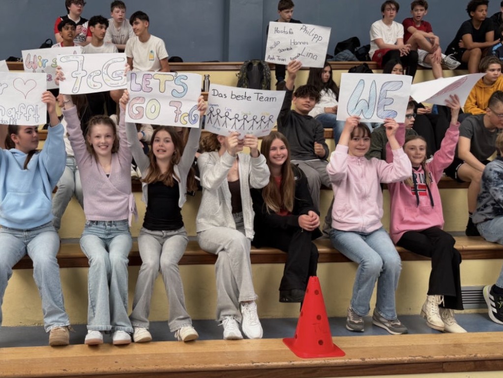 Jugendliche sitzen auf den Rängen einer Sporthalle und halten selbst gebastelte Plakate hoch, um ihre Klassenmannschaft anzufeuern.