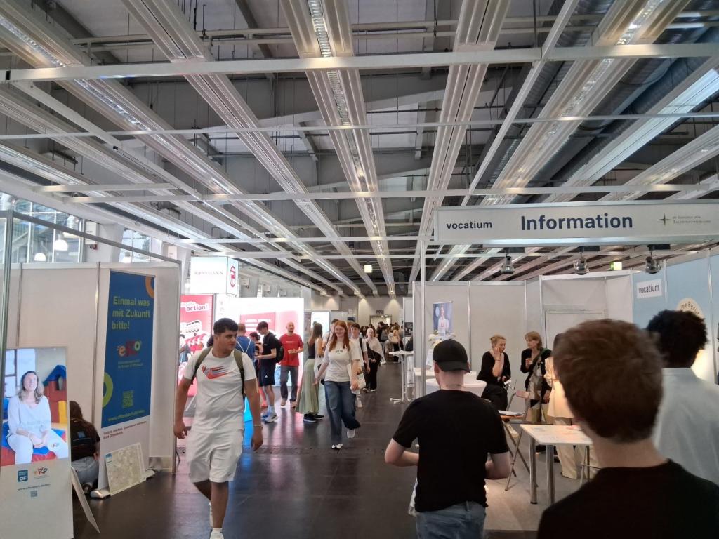 Blick in eine große Messehalle mit Ausstellungsständen und einem Schild „vocatium Information“; im Gang gehen Jugendliche und Besucher zwischen den Ständen entlang.