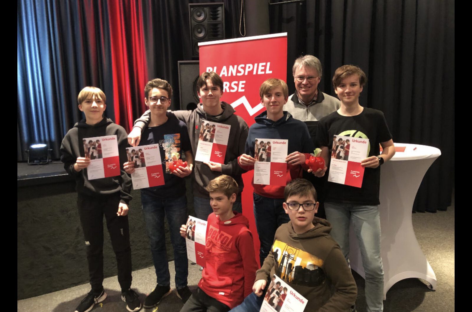 Eine Gruppe Jugendlicher hält Urkunden in die Kamera; hinter ihnen ein rotes Roll-up mit der Aufschrift „Planspiel Börse“, daneben eine erwachsene Begleitperson.