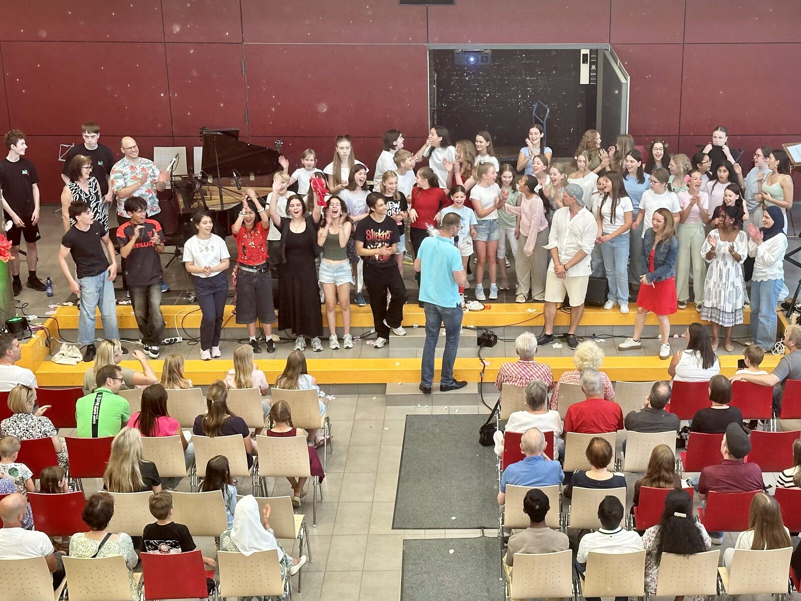 Eine große Gruppe Jugendlicher und Erwachsener steht nach einem Auftritt jubelnd auf dem Podium der Aula, neben einem Flügel; davor sitzt das Publikum in Stuhlreihen.