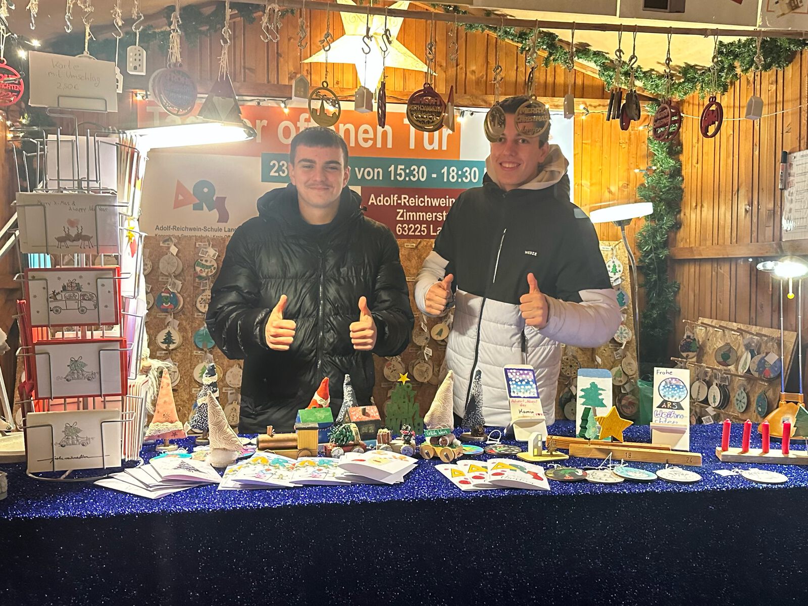 Zwei Jugendliche stehen mit erhobenen Daumen hinter einem weihnachtlich geschmückten Verkaufsstand mit selbst gebastelten Karten, Holzanhängern und Deko; im Hintergrund ein Banner der Adolf-Reichwein-Schule Langen.