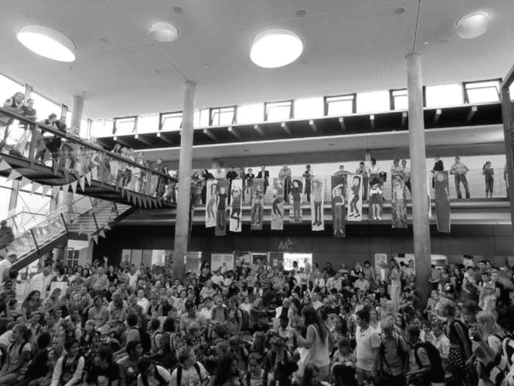 Das Bild zeigt eine große Menschenmenge in der Aula. Die Menschen stehen auf einer Treppe und in einem Bereich darunter. Es gibt auch einige Banner an der Wand. Das Bild ist in Schwarzweiß gehalten und die Eltern und die neuen fünft Klässler nehmen an der Einschulung teil.