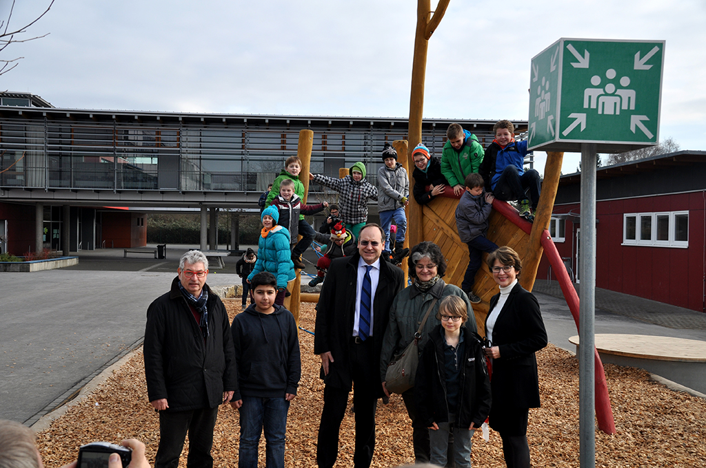 Der neue Schulhof wo Kinder drauf Klettern und Erwachsene davor stehen Das Bild zeigt eine Gruppe von Schülern, die auf den neuen Hof stehen. Einige der Kinder klettern auf einem Holzgerüst und andere stehen unten. Es sind auch Erwachsene wie die Schulleiterin auf dem Bild zu sehen wie die Schulleiterin . Im Hintergrund ist das B Gebäude zu sehen zu sehen.