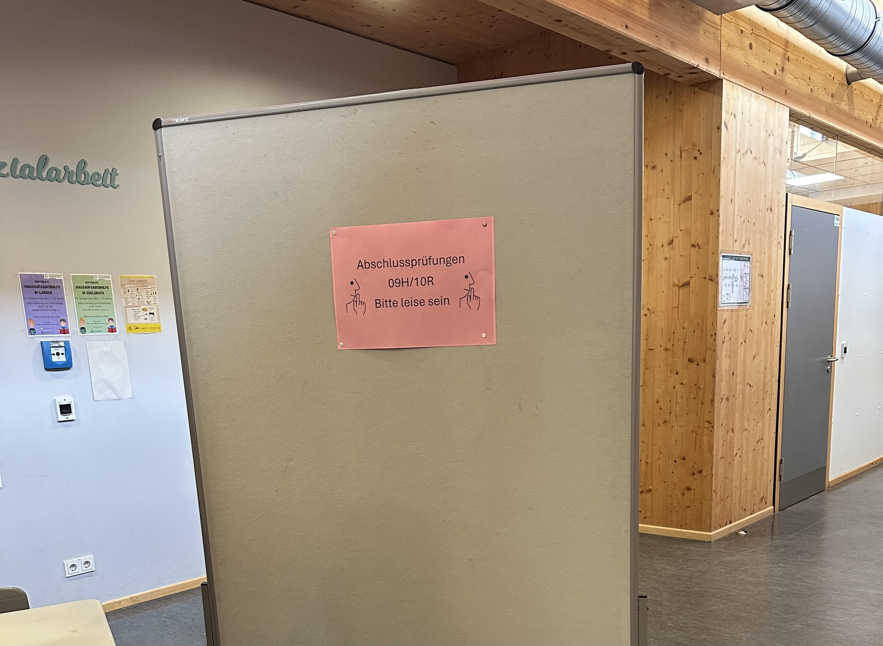Das Foto zeigt einen ruhigen Schulflur einer Realschule. Im Mittelpunkt steht eine große, freistehende graue Stellwand, die fast den gesamten Blick versperrt. An der Stellwand hängt ein rosa Blatt Papier mit der Aufschrift:    „Abschlussprüfungen 09H/10R – Bitte leise sein“    Neben dem Text sind auf beiden Seiten Symbole von Fingern zu sehen, die zum Stillsein auffordern.    Der Flur ist modern gestaltet. Rechts sind Wände und Stützen aus hellem Holz zu sehen, darüber verläuft ein silbernes Lüftungsrohr unter der Decke. Im Hintergrund führt ein Gang zu mehreren Räumen mit geschlossenen Türen.